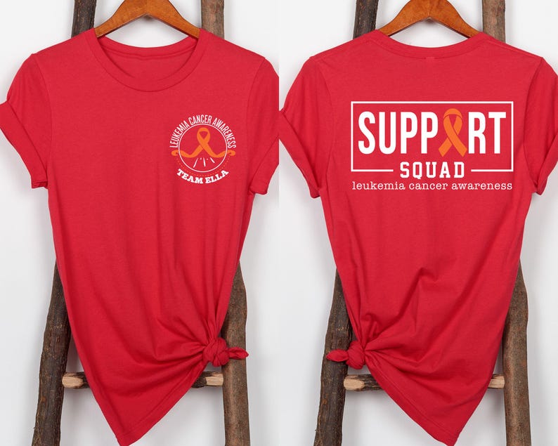 Puede incluir: Una camiseta roja con un gr&aacute;fico frontal que muestra una cinta naranja y el texto "LEUKEMIA CANCER AWARENESS TEAM ELLA". El gr&aacute;fico trasero muestra un marco rectangular blanco con el texto "SUPPORT SQUAD" y "leukemia cancer awareness".