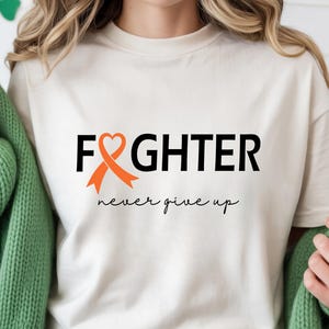 Puede incluir: Una camiseta de color crema con el texto negro "FIGHTER" y un lazo de concienciaci&oacute;n naranja con forma de coraz&oacute;n que sustituye a la letra "I". Debajo del texto principal, la frase "never give up" est&aacute; impresa en una fuente cursiva negra.