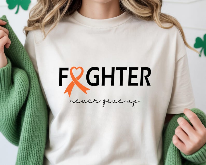 Puede incluir: Una camiseta de color crema con el texto negro "FIGHTER" y un lazo de concienciaci&oacute;n naranja con forma de coraz&oacute;n que sustituye a la letra "I". Debajo del texto principal, la frase "never give up" est&aacute; impresa en una fuente cursiva negra.