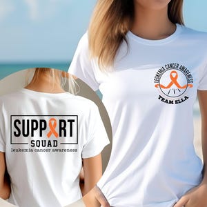 Puede incluir: Una camiseta blanca con un dise&ntilde;o de concienciaci&oacute;n sobre la leucemia. El pecho muestra una cinta naranja con el texto LEUKEMIA CANCER AWARENESS TEAM ELLA. La espalda presenta un gr&aacute;fico rectangular con SUPPORT SQUAD y leukemia cancer awareness.