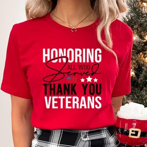 May include: Red t-shirt with white text that reads "Honoring All Who Served Thank You Veterans". The text is in a bold, cursive font. There is a small American flag graphic and five stars above the word "Served".