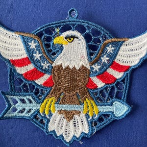 May include: Embroidered patch featuring a detailed bald eagle design. The eagle has white and brown feathers, a yellow beak and talons, and is set against a backdrop of red, white, and blue. The patch includes an arrow and a loop for hanging.
