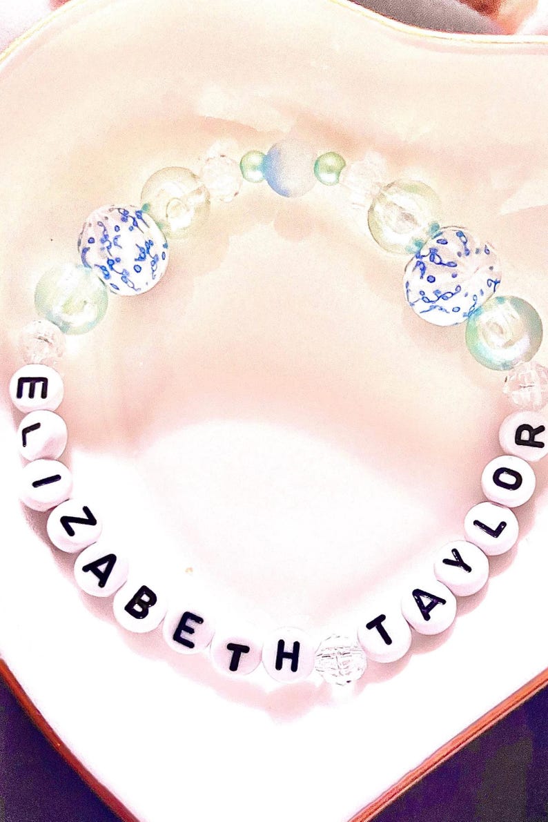 collage featuring two handmade Taylor Swift The Life of a Showgirl inspired friendship bracelets. One bracelet is shimmering blue with letter beads spelling “Elizabeth Taylor,” capturing the timeless glamor of the track. The other bracelet is soft pink with blue hues, spelling “Ophelia,” inspired by the poetic song The Fate of Ophelia. Both elastic beaded bracelets are perfect Swiftie jewelry, concert accessories, or collectible fan gifts from Taylor’s newest album era