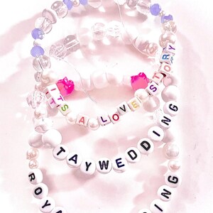 May include: Three elastic beaded bracelets featuring white, clear, and purple beads. One bracelet displays the text IT'S A LOVE STORY with pink heart beads, another reads TAYWEDDING, and the third features the text ROYAL WEDDING in black letters.