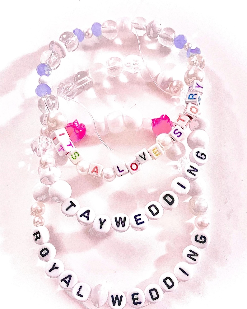 May include: Three elastic beaded bracelets featuring white, clear, and purple beads. One bracelet displays the text IT'S A LOVE STORY with pink heart beads, another reads TAYWEDDING, and the third features the text ROYAL WEDDING in black letters.