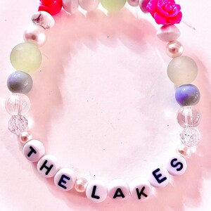 K&ouml;nnte beinhalten: Ein Perlenarmband mit wei&szlig;en runden Perlen, die den schwarzen Schriftzug THE LAKES tragen, durchsetzt mit verschiedenen dekorativen Elementen, darunter zwei leuchtend pinke rosenf&ouml;rmige Perlen, transparente klare Perlen, mattierte hellgr&uuml;ne Kugeln und wei&szlig;e Perlen.
