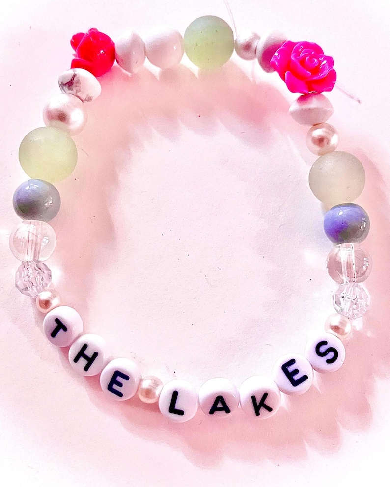 K&ouml;nnte beinhalten: Ein Perlenarmband mit wei&szlig;en runden Perlen, die den schwarzen Schriftzug THE LAKES tragen, durchsetzt mit verschiedenen dekorativen Elementen, darunter zwei leuchtend pinke rosenf&ouml;rmige Perlen, transparente klare Perlen, mattierte hellgr&uuml;ne Kugeln und wei&szlig;e Perlen.