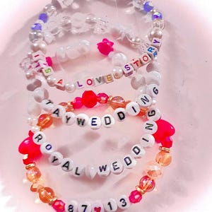 May include: Three beaded bracelets featuring white letter beads spelling out IT'S A LOVE STORY, TAY WEDDING, and ROYAL WEDDING 87 13. The bracelets include clear, iridescent, orange, and hot pink beads, with small heart-shaped accents interspersed.