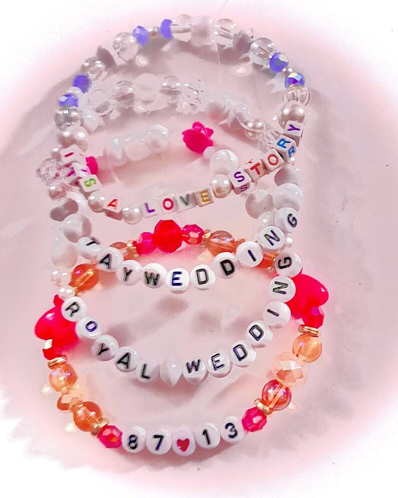 May include: Three beaded bracelets featuring white letter beads spelling out IT'S A LOVE STORY, TAY WEDDING, and ROYAL WEDDING 87 13. The bracelets include clear, iridescent, orange, and hot pink beads, with small heart-shaped accents interspersed.