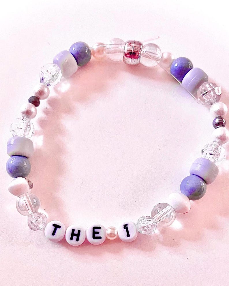 Handmade “The 1” friendship bracelet inspired by Taylor Swift’s Folklore album. Features white, grey, and silver beads with white howlite stone accents and “The 1” letter beads on elastic stretch cord. Created with a mix of glass, plastic, and whenever possible, upcycled and thrifted beads. Perfect for Swifties, Folklore fans, friendship bracelet collectors, concert outfits, bracelet stacks, and unique handmade gifts.
