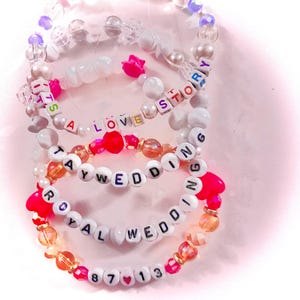 May include: A set of four elastic beaded bracelets featuring white letter beads spelling out IT'S A LOVE STORY, TAY WEDDING, ROYAL WEDDING, and 87 13. The bracelets include clear, white, orange, and pink beads with small pink heart-shaped accents.
