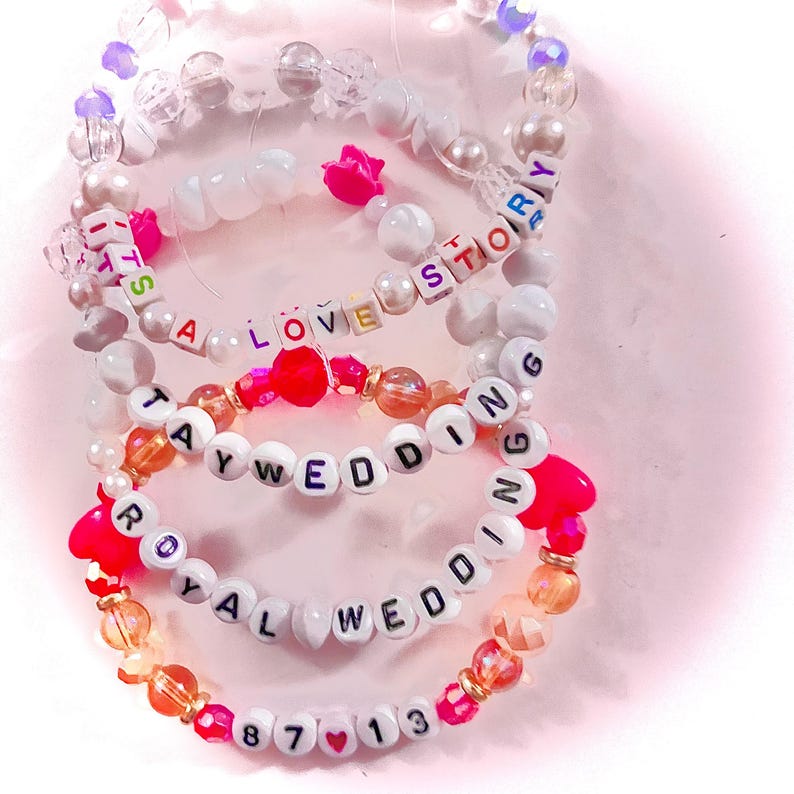 May include: A set of four elastic beaded bracelets featuring white letter beads spelling out IT'S A LOVE STORY, TAY WEDDING, ROYAL WEDDING, and 87 13. The bracelets include clear, white, orange, and pink beads with small pink heart-shaped accents.