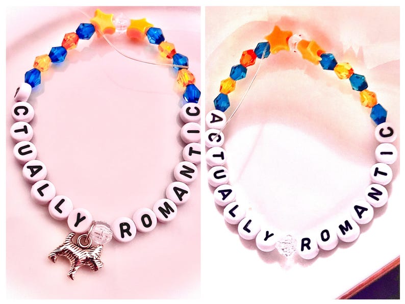 collage showcasing two handmade Taylor Swift The Life of a Showgirl inspired friendship bracelets. Both feature the phrase “Actually Romantic” in letter beads, celebrating the sweet and dreamy track from the album. One bracelet is designed in bold orange and green/teal beads, while the other is crafted in soft pink tones. Made with stretchy elastic, glass, and plastic beads, these Swiftie bracelets are perfect jewelry gifts, concert accessories, or collectible fan merch