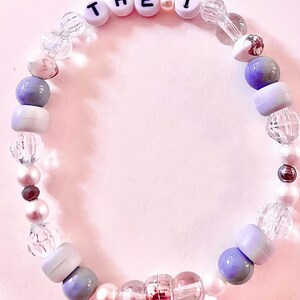 K&ouml;nnte beinhalten: Ein kreisf&ouml;rmiges Perlenarmband mit einer Mischung aus klaren facettierten Perlen, glatten wei&szlig;en Perlen sowie matten violetten und grauen Perlen. Das Armband enth&auml;lt wei&szlig;e Buchstabenperlen mit der Aufschrift THE 1, unterbrochen von kleinen wei&szlig;en Zwischenperlen.