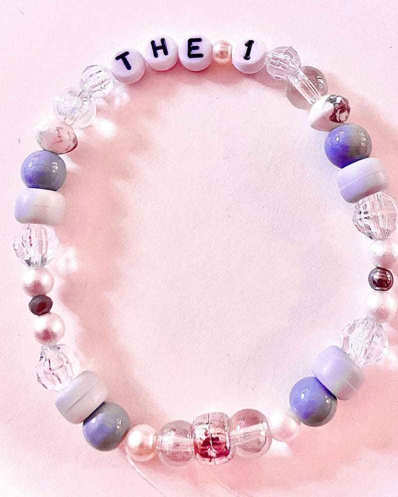 K&ouml;nnte beinhalten: Ein kreisf&ouml;rmiges Perlenarmband mit einer Mischung aus klaren facettierten Perlen, glatten wei&szlig;en Perlen sowie matten violetten und grauen Perlen. Das Armband enth&auml;lt wei&szlig;e Buchstabenperlen mit der Aufschrift THE 1, unterbrochen von kleinen wei&szlig;en Zwischenperlen.