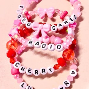 May include: A set of four beaded bracelets featuring pink and red translucent, opaque, and faceted beads. The bracelets include pink plastic bow charms, star-shaped beads, and white letter beads spelling out VIDEO GAMES, RADIO, CHERRY, and LUST FOR.