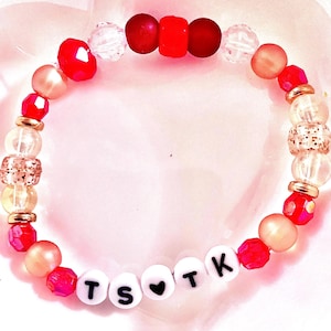 May include: A beaded bracelet featuring a mix of red, clear, and metallic gold beads. The bracelet includes white round letter beads spelling out TS heart TK, with a black heart symbol between the letters. The design incorporates faceted, smooth, and textured beads arranged in a circular pattern.