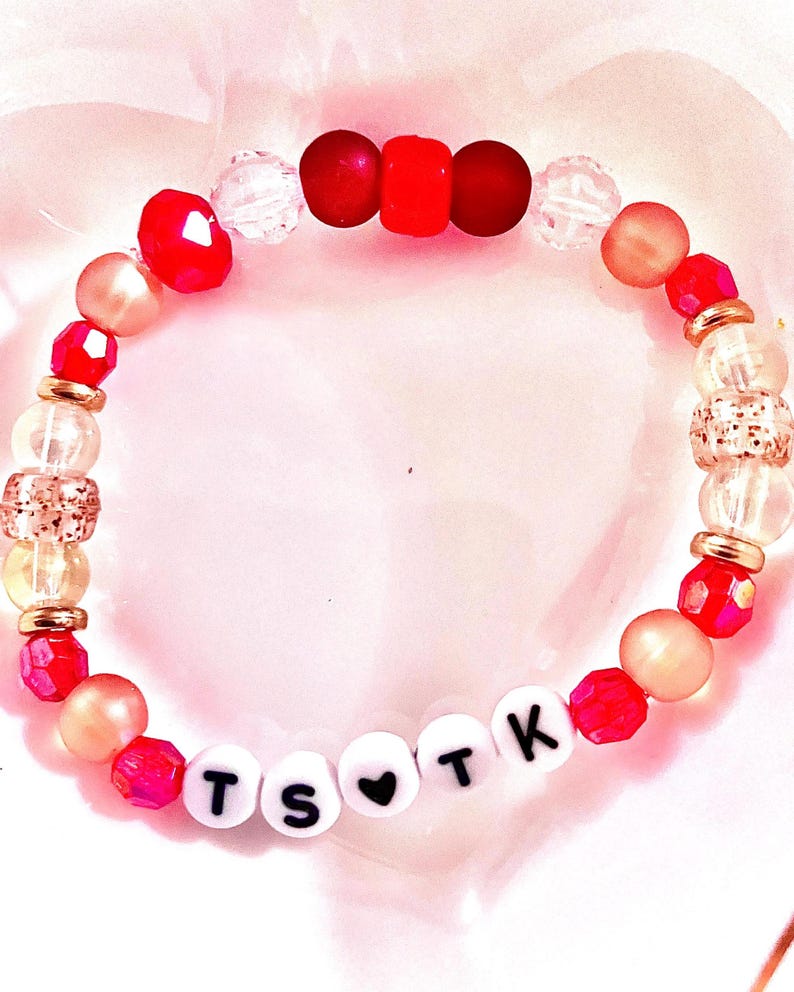 May include: A beaded bracelet featuring a mix of red, clear, and metallic gold beads. The bracelet includes white round letter beads spelling out TS heart TK, with a black heart symbol between the letters. The design incorporates faceted, smooth, and textured beads arranged in a circular pattern.