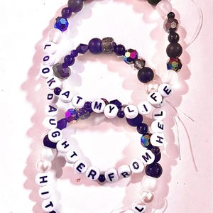 May include: Two elastic bracelets featuring white letter beads spelling LOOK AT MY LIFE DAUGHTER FROM HELL HIT THE WALL. The bracelets include a mix of matte black, translucent white, and iridescent faceted beads in shades of purple and dark blue.