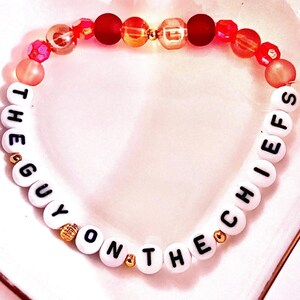 May include: A beaded bracelet featuring white round beads with black letters spelling out THE GUY ON THE CHIEFS, interspersed with smaller gold-toned metallic beads. The upper section consists of a mix of red, orange, and translucent pink beads.