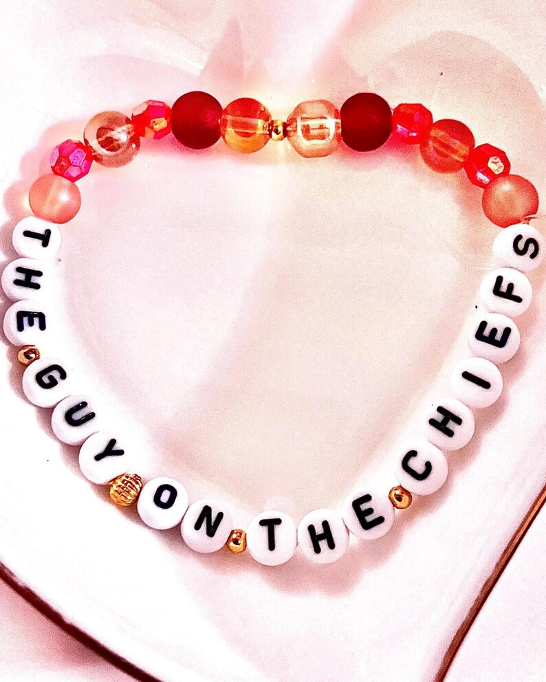 May include: A beaded bracelet featuring white round beads with black letters spelling out THE GUY ON THE CHIEFS, interspersed with smaller gold-toned metallic beads. The upper section consists of a mix of red, orange, and translucent pink beads.