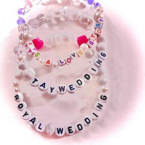 May include: Three beaded bracelets featuring white, clear, and purple beads. One bracelet displays the text IT'S A LOVE STORY, another TAYWEDDING, and the third ROYAL WEDDING. The bracelets include small pink heart-shaped beads and round white pearls.