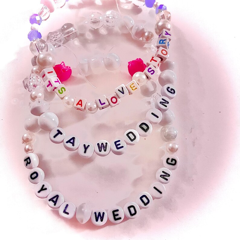 May include: Three beaded bracelets featuring white, clear, and purple beads. One bracelet displays the text IT'S A LOVE STORY, another TAYWEDDING, and the third ROYAL WEDDING. The bracelets include small pink heart-shaped beads and round white pearls.
