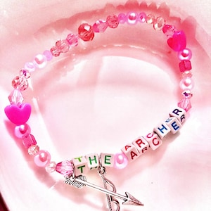 This pink glass and acrylic beaded bracelet, inspired by Taylor Swift's "The Archer," showcases a bow and arrow charm, symbolizing the song's themes of vulnerability and self-discovery. A must-have for dedicated Swifties in their Lover era.