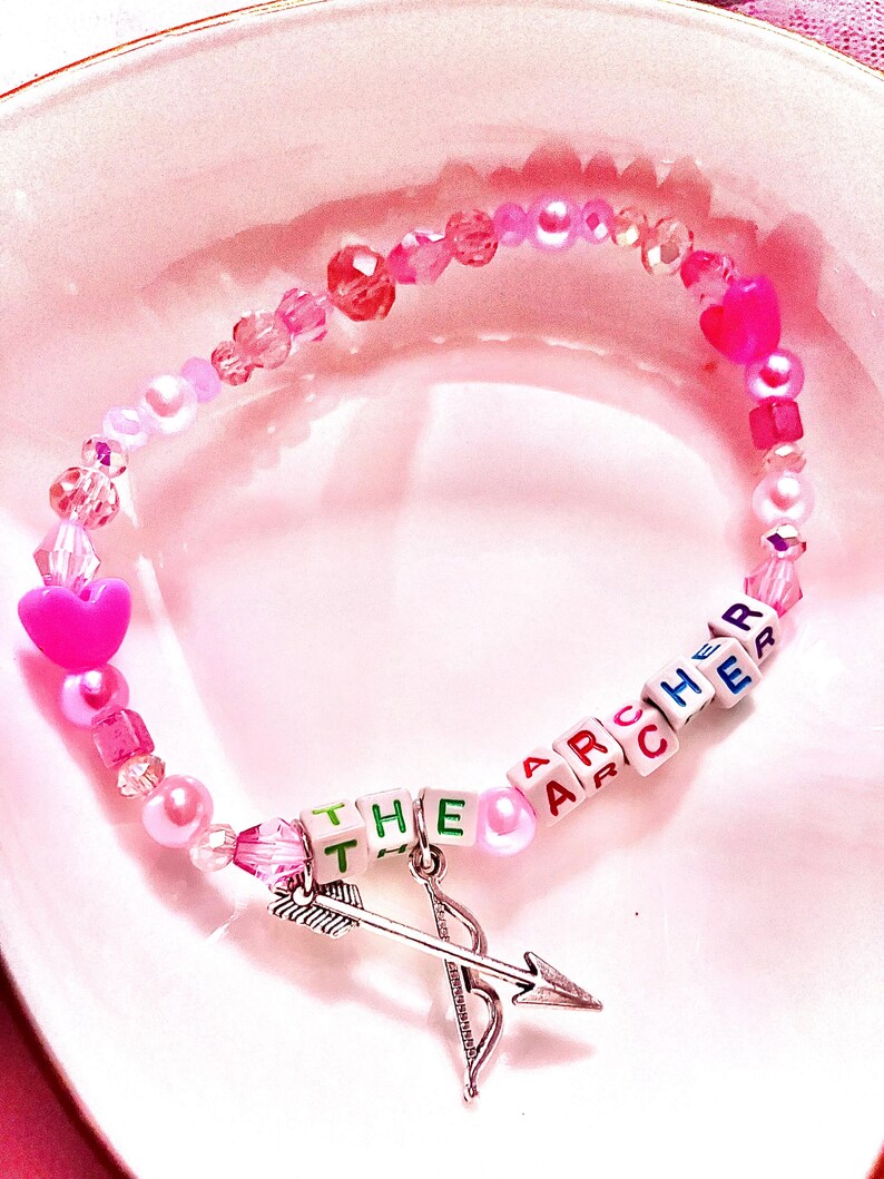This pink glass and acrylic beaded bracelet, inspired by Taylor Swift's "The Archer," showcases a bow and arrow charm, symbolizing the song's themes of vulnerability and self-discovery. A must-have for dedicated Swifties in their Lover era.