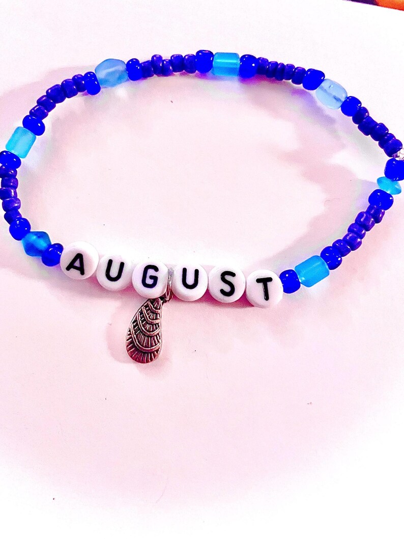 K&ouml;nnte beinhalten: Ein Perlenarmband mit einer Mischung aus dunkelblauen und hellblauen Perlen. In der Mitte befinden sich wei&szlig;e, runde Perlen mit dem schwarzen Schriftzug AUGUST. Ein kleiner, silberfarbener Metallanh&auml;nger in Muschelform h&auml;ngt in der Mitte.