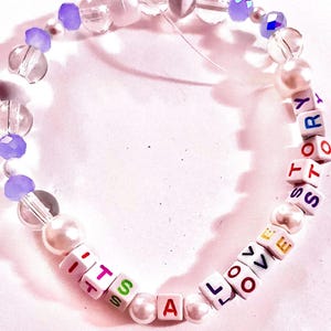 May include: A beaded bracelet featuring white square letter beads spelling out the phrase ITS A LOVE STORY in multicolored text. The design includes clear faceted beads, opaque white pearl-like beads, and translucent purple accent beads.