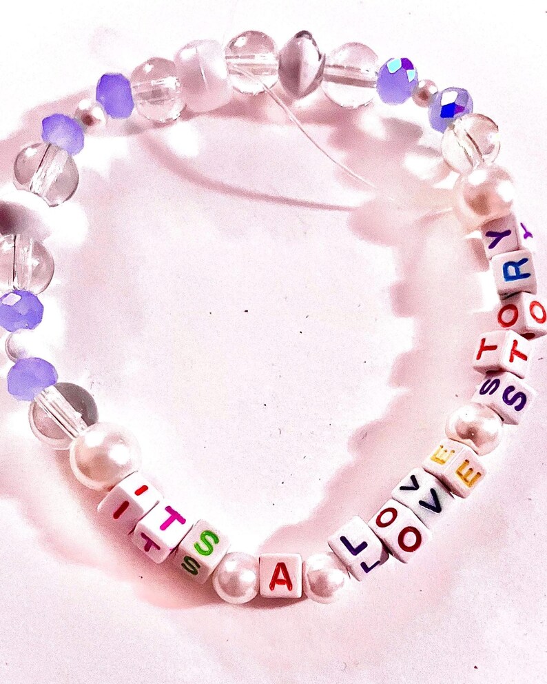 May include: A beaded bracelet featuring white square letter beads spelling out the phrase ITS A LOVE STORY in multicolored text. The design includes clear faceted beads, opaque white pearl-like beads, and translucent purple accent beads.