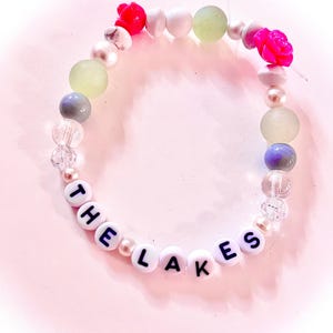 Handmade “The Lakes” friendship bracelet inspired by Taylor Swift’s Folklore album. Features white, green, and pink beads with “The Lakes” letter beads and two pink rose accents on elastic stretch cord. Made with a mix of glass, plastic, and whenever possible, upcycled and thrifted beads. Cottagecore inspired bracelet perfect for Swifties, Folklore fans, friendship bracelet collectors, concert outfits, bracelet stacks, and unique handmade gifts.