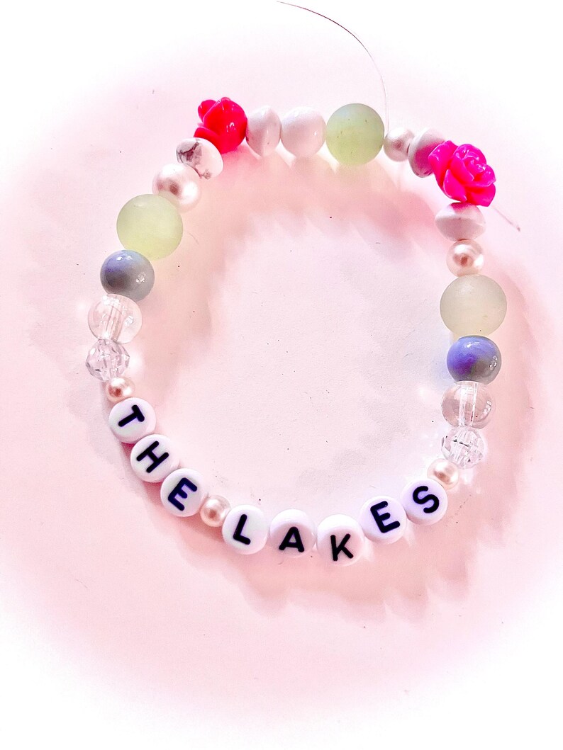 Handmade “The Lakes” friendship bracelet inspired by Taylor Swift’s Folklore album. Features white, green, and pink beads with “The Lakes” letter beads and two pink rose accents on elastic stretch cord. Made with a mix of glass, plastic, and whenever possible, upcycled and thrifted beads. Cottagecore inspired bracelet perfect for Swifties, Folklore fans, friendship bracelet collectors, concert outfits, bracelet stacks, and unique handmade gifts.