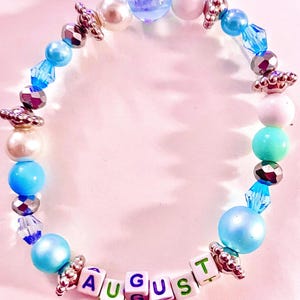 K&ouml;nnte beinhalten: Ein Perlenarmband mit einer Vielzahl von blauen, wei&szlig;en und silberfarbenen Perlen, darunter runde Perlen, facettierte Kristalle und metallische Zwischenst&uuml;cke. Das Armband enth&auml;lt wei&szlig;e quadratische Buchstabenperlen, die das Wort AUGUST in Lila und Gr&uuml;n bilden.