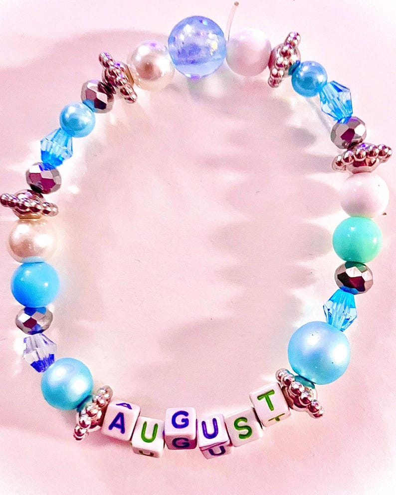 K&ouml;nnte beinhalten: Ein Perlenarmband mit einer Vielzahl von blauen, wei&szlig;en und silberfarbenen Perlen, darunter runde Perlen, facettierte Kristalle und metallische Zwischenst&uuml;cke. Das Armband enth&auml;lt wei&szlig;e quadratische Buchstabenperlen, die das Wort AUGUST in Lila und Gr&uuml;n bilden.