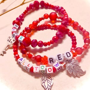May include: Three stacked beaded bracelets in shades of red, featuring faceted, matte, and smooth round beads. White square letter beads spell out "BABE", "ALL TOO WELL", and "RED". Silver-toned metal charms include a key and two leaf shapes.