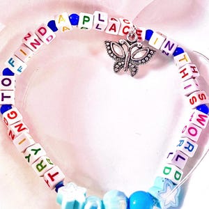 May include: A circular bracelet made of white square beads with colorful letters spelling out TRYING TO FIND A PLACE IN THIS WORLD. The bracelet features a silver butterfly charm, small blue round beads, and light blue star-shaped beads.