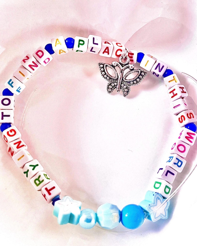 May include: A circular bracelet made of white square beads with colorful letters spelling out TRYING TO FIND A PLACE IN THIS WORLD. The bracelet features a silver butterfly charm, small blue round beads, and light blue star-shaped beads.