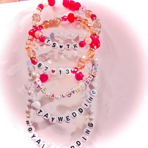 May include: A stack of five beaded bracelets featuring colorful translucent, opaque, and pearl-like beads. The bracelets include white letter beads spelling out TS TK, 87 13, IT'S A LOVE STORY, TAYWEDDING, and ROYAL WEDDING, with small heart-shaped beads.