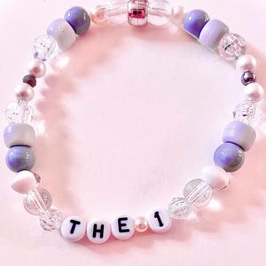 K&ouml;nnte beinhalten: Ein Perlenarmband mit einer Mischung aus violetten, wei&szlig;en und klaren facettierten Perlen. Das Armband enth&auml;lt wei&szlig;e Buchstabenperlen, die den Text THE 1 bilden, durchsetzt mit kleinen perlmuttartigen Perlen und einer silberfarbenen Mittelperle.
