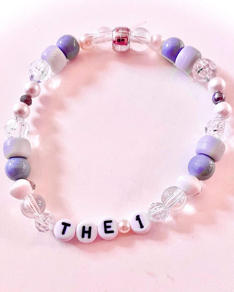 K&ouml;nnte beinhalten: Ein Perlenarmband mit einer Mischung aus violetten, wei&szlig;en und klaren facettierten Perlen. Das Armband enth&auml;lt wei&szlig;e Buchstabenperlen, die den Text THE 1 bilden, durchsetzt mit kleinen perlmuttartigen Perlen und einer silberfarbenen Mittelperle.