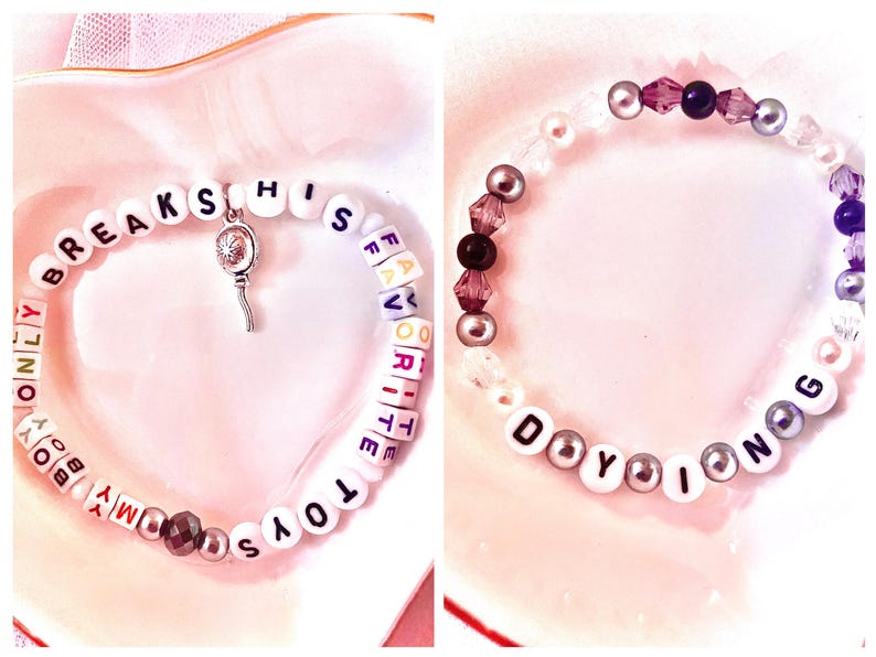 May include: Two beaded bracelets. The first bracelet is white with black lettering spelling "My Boy Only Breaks His Favourite Toys". The second bracelet is white with black lettering spelling "Do You Know G" and has purple and silver beads.