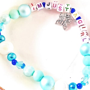May include: A beaded bracelet featuring light blue and white pearls, translucent blue crystals, and white square letter beads spelling out I M JUST A GIRL. A silver-toned metal butterfly charm hangs from the center of the beaded strand.
