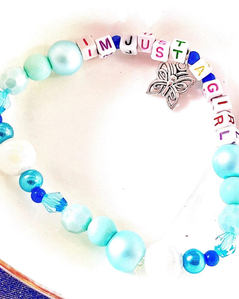 May include: A beaded bracelet featuring light blue and white pearls, translucent blue crystals, and white square letter beads spelling out I M JUST A GIRL. A silver-toned metal butterfly charm hangs from the center of the beaded strand.