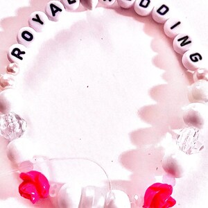 White and pink handmade “royal wedding” friendship bracelet inspired by Taylor Swift and Travis Kelce with soft romantic bead details and two pink rose accents. Elegant elastic stretch bracelet inspired by celebrity romance, garden engagement aesthetics, and American royalty style. Perfect for Swiftie collections, feminine bracelet stacks, concert accessories, friendship bracelet trading, and romantic pop culture jewelry.