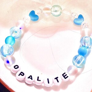 A handmade Taylor Swift The Life of a Showgirl inspired friendship bracelet featuring the track “Opalite.” This bracelet is crafted with shimmering opalite beads and soft blue accents, strung on durable elastic with letter beads spelling “Opalite.” A dreamy, crystal-inspired design perfect for Swifties, crystal lovers, and fans of Taylor’s newest album era. Trendy beaded bracelet for stacking, gifting, or wearing to concerts as a unique piece of Swiftie jewelry
