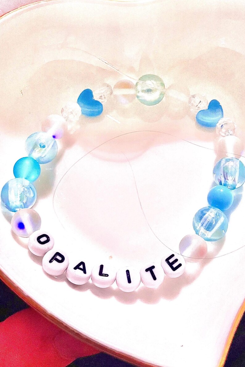 A handmade Taylor Swift The Life of a Showgirl inspired friendship bracelet featuring the track “Opalite.” This bracelet is crafted with shimmering opalite beads and soft blue accents, strung on durable elastic with letter beads spelling “Opalite.” A dreamy, crystal-inspired design perfect for Swifties, crystal lovers, and fans of Taylor’s newest album era. Trendy beaded bracelet for stacking, gifting, or wearing to concerts as a unique piece of Swiftie jewelry