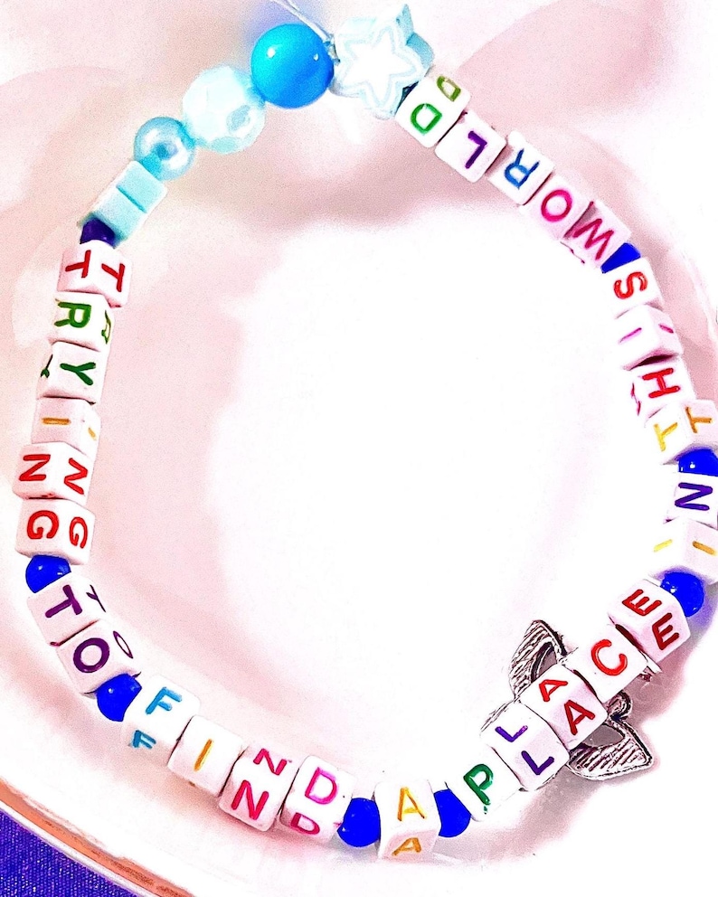 May include: A beaded bracelet featuring white square letter beads spelling out the phrase TRYING TO FIND A PLACE IN THIS WORLD in multicolored text. The bracelet includes blue round beads, a star-shaped bead, and a silver-toned metal charm.