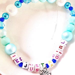 May include: A beaded bracelet featuring a mix of light blue, turquoise, and white round beads, interspersed with small dark blue beads. White square letter beads spell out the phrase I AM JUST A GIRL, accented by a silver-toned butterfly charm.
